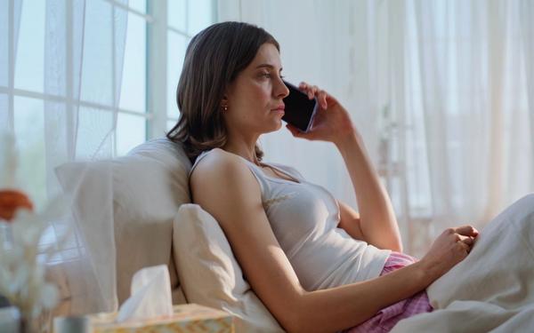 A sick woman in bed holds a phone to her ear with a thermometer under her arm, calling off work on short notice.