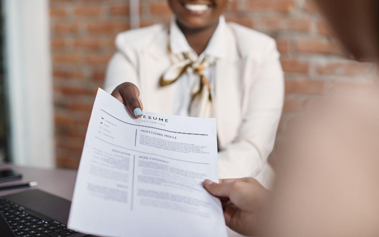 A job applicant smiling as they hand a resume to an interviewer, with the resume displaying a professional bio.