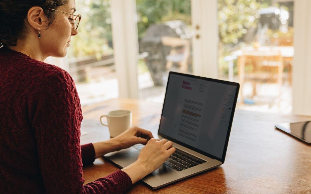 Person sitting at a wooden table in a bright room, typing on a laptop with a cover letter visible on the screen, representing someone writing or editing a professional cover letter.