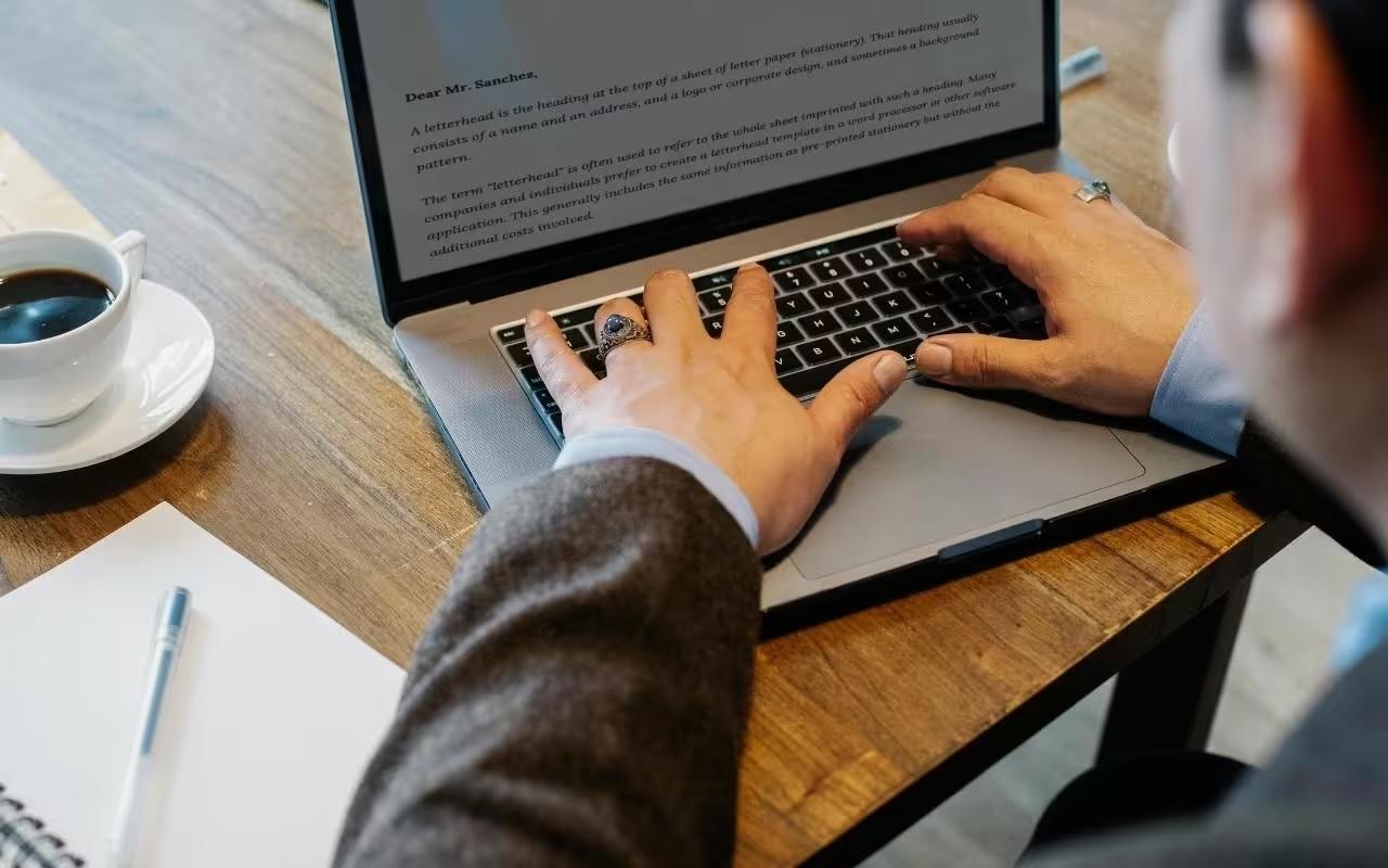 Over-the-shoulder view of someone writing a business letter on a laptop next to a notebook and a cup of coffee.