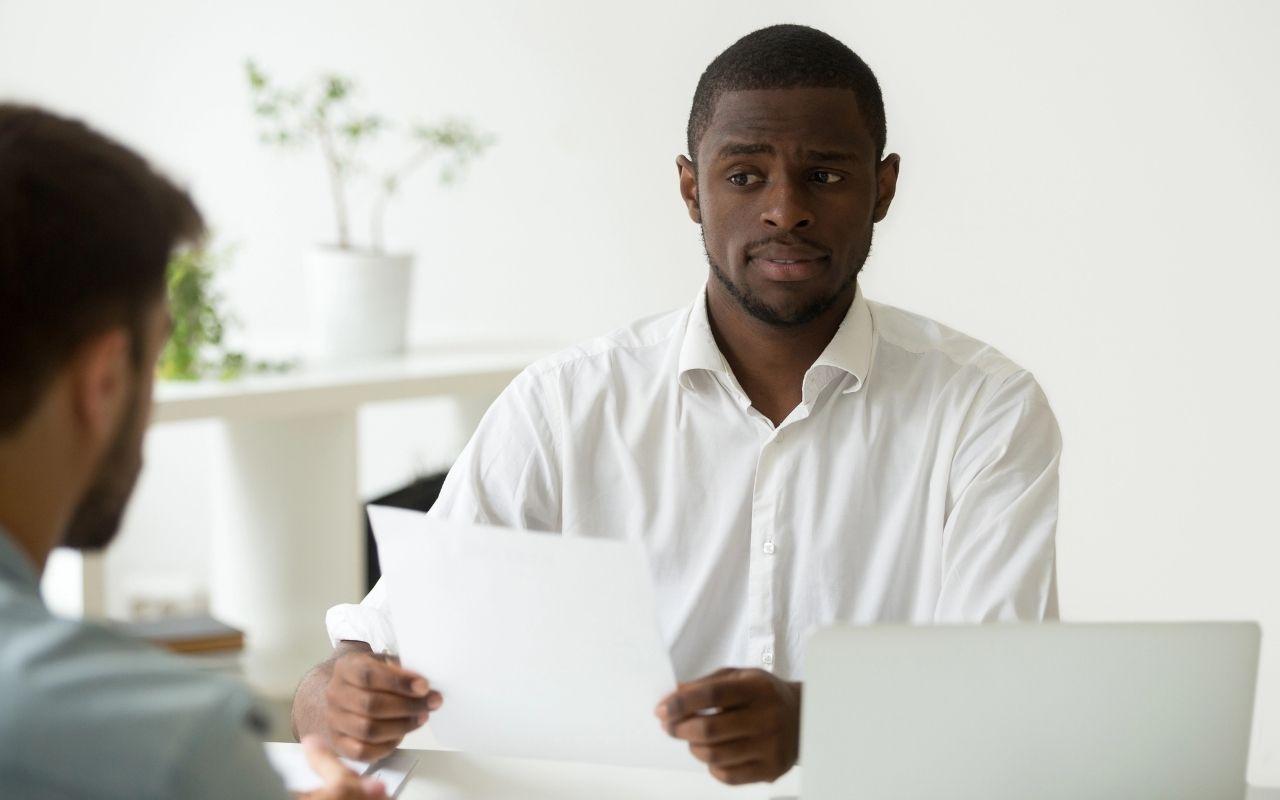 A hiring manager looks suspiciously at a candidate, catching a lie or discrepancy on a printed resume.