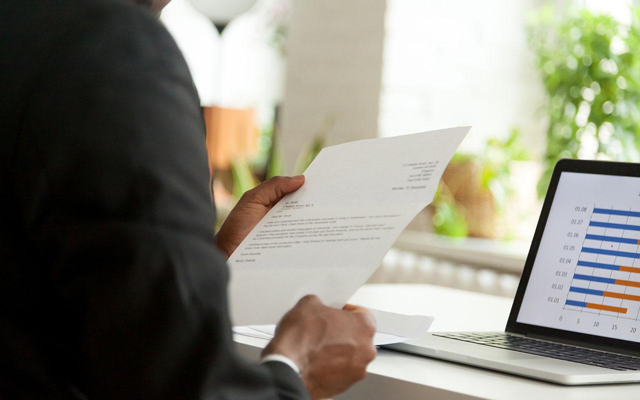 A person in business attire reads a cover letter at their desk, with a laptop displaying a bar chart beside them.