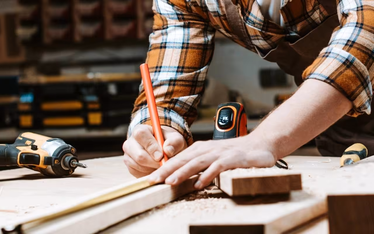 Close-up of a carpenter using a pencil and a tape measure to mark a piece of wood in a workshop, representing hands-on jobs.