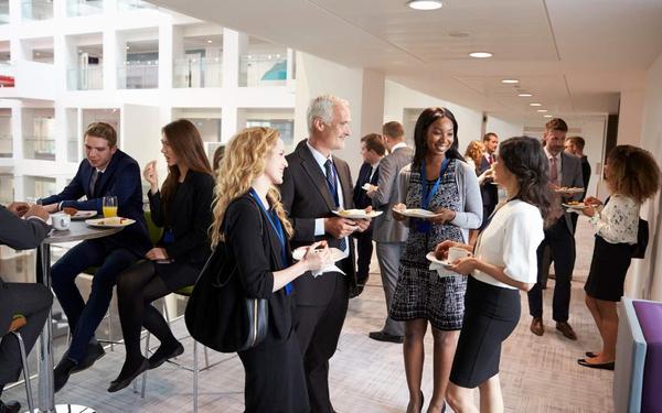 Diverse business professionals network, chat, and eat food at a corporate event in a bright, modern office.
