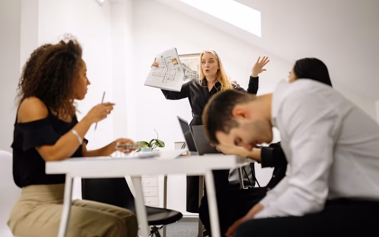 An angry boss gestures wildly with a document at a meeting while a stressed employee slumps forward in despair.