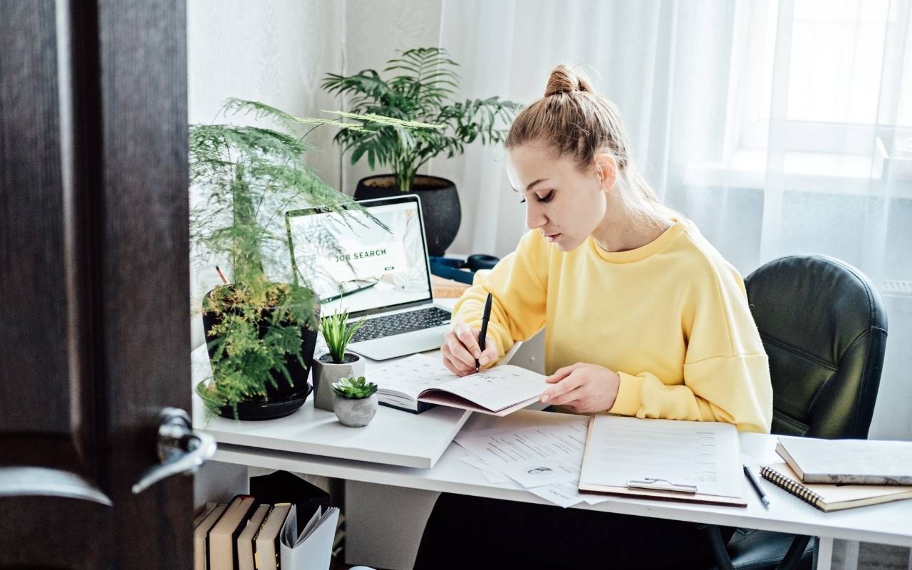 Person writing in a planner at a desk with a laptop displaying a job search, representing career path evaluation.