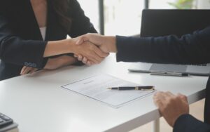 Two people shake hands across a desk during a job interview. On the desk between them lies a cover letter.