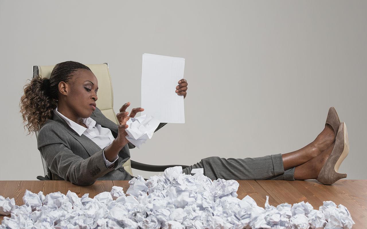 A businesswoman leans back with her feet on a desk piled with crumpled papers, looking frustrated while reviewing a document, illustrating bad resumes that get rejected by hiring managers.