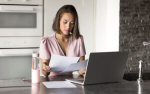 Woman sitting at a kitchen counter reviewing documents while working on a laptop, representing writing a cover letter and resume.