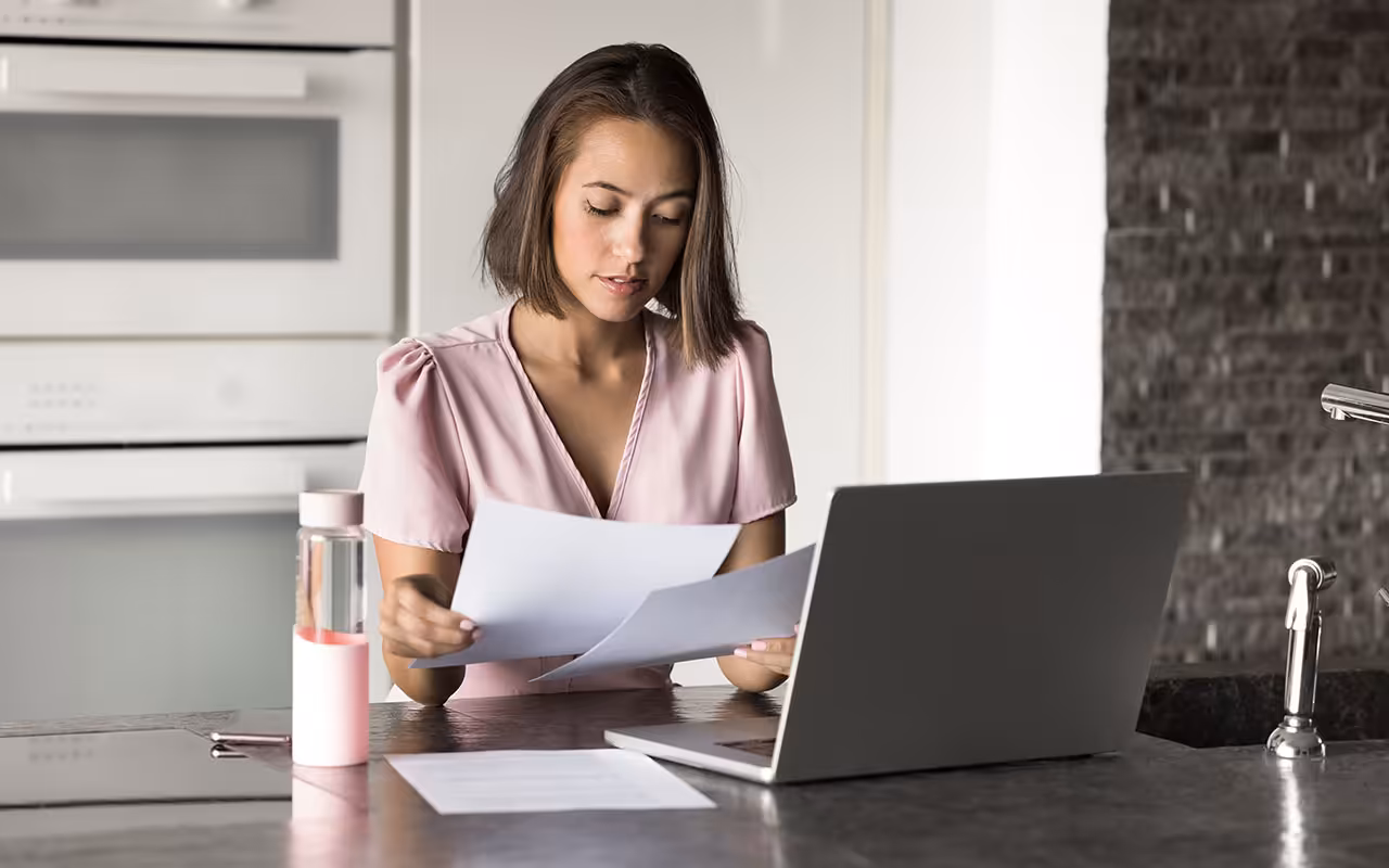 Woman sitting at a kitchen counter reviewing documents while working on a laptop, representing writing a cover letter and resume.
