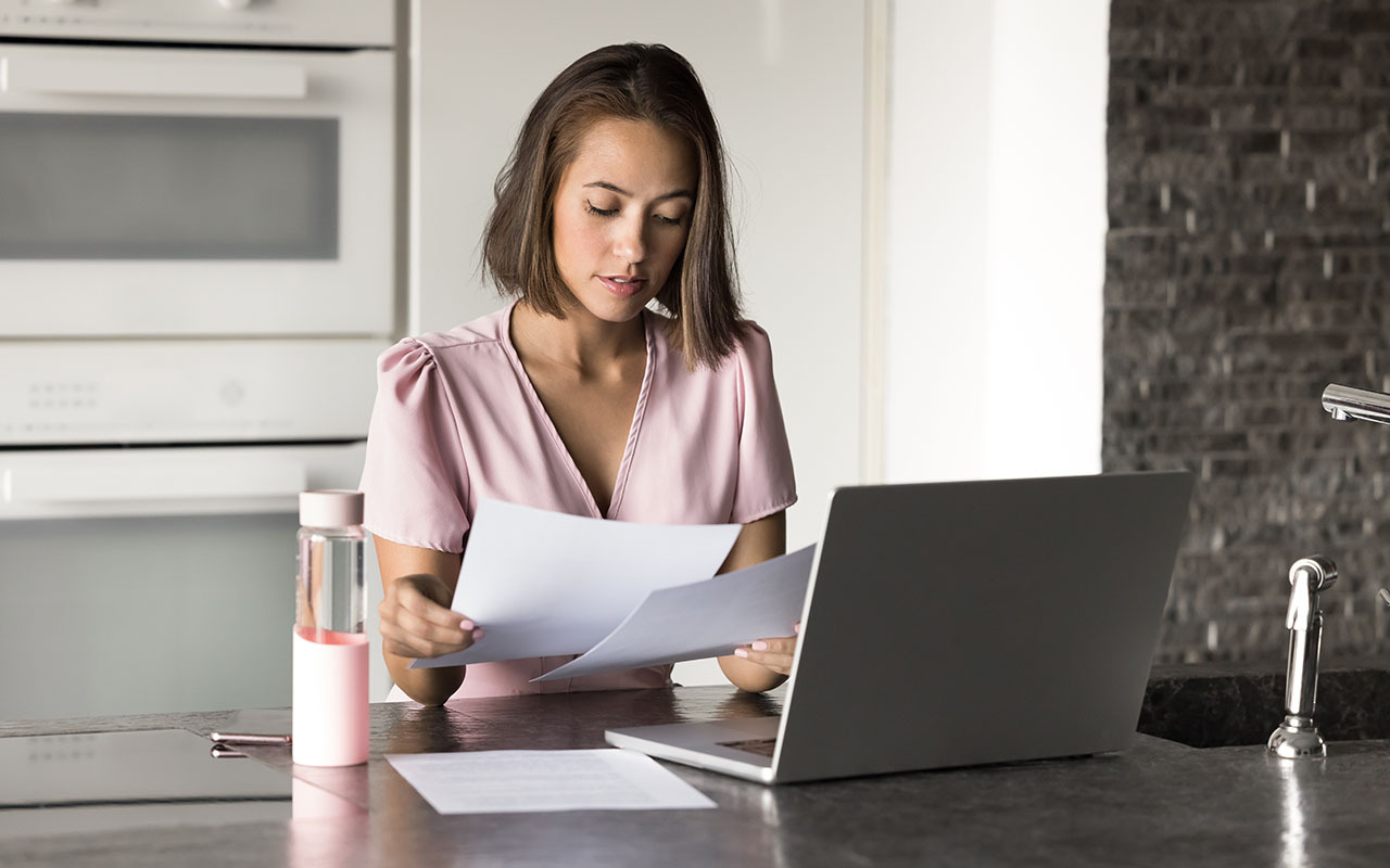 Woman sitting at a kitchen counter reviewing documents while working on a laptop, representing writing a cover letter and resume.