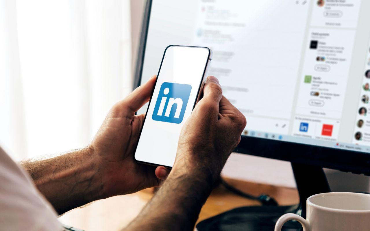 A person holds a phone with the LinkedIn logo next to a computer screen showing LinkedIn, illustrating how to add a resume.