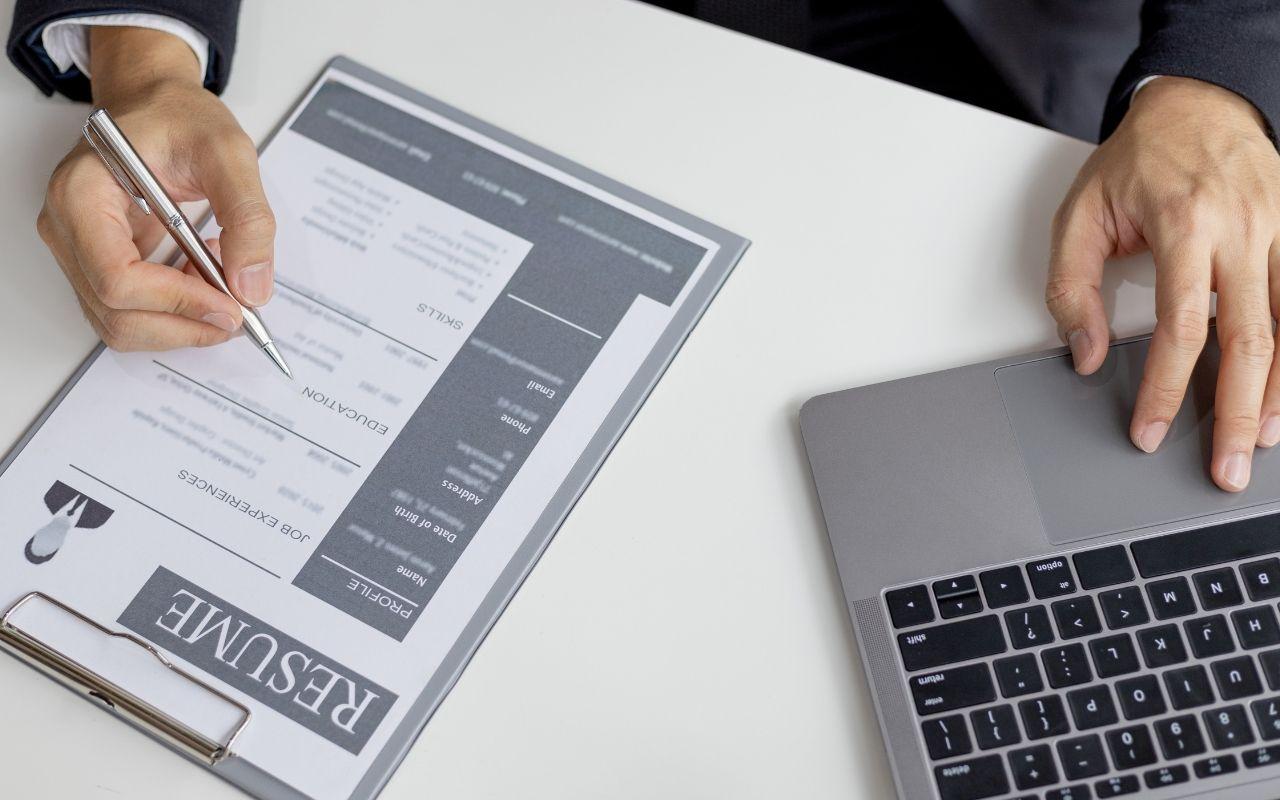 Top-down view of a person using a laptop and pointing a pen at the education section of a resume on a desk.