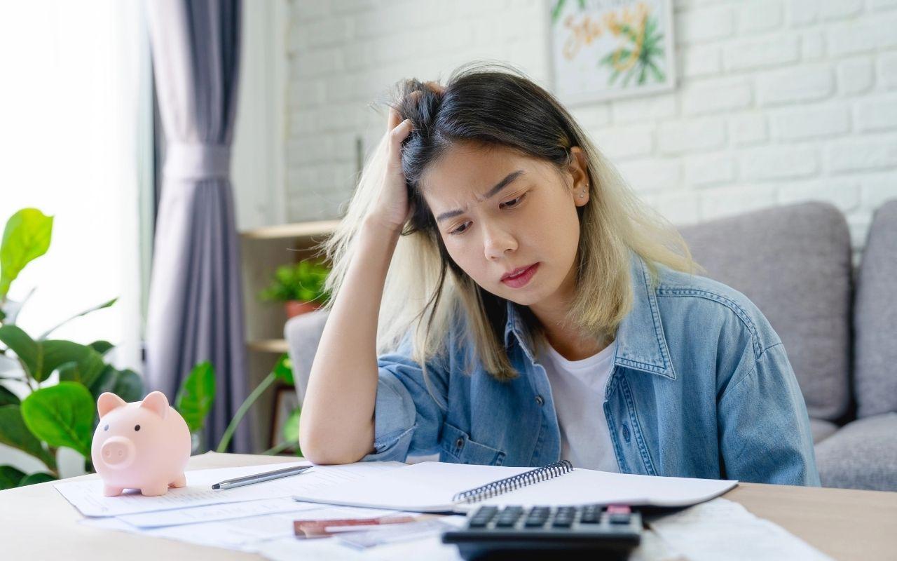 A worried young woman reviews finances at home, looking stressed with a notebook, calculator, and a pink piggy bank.