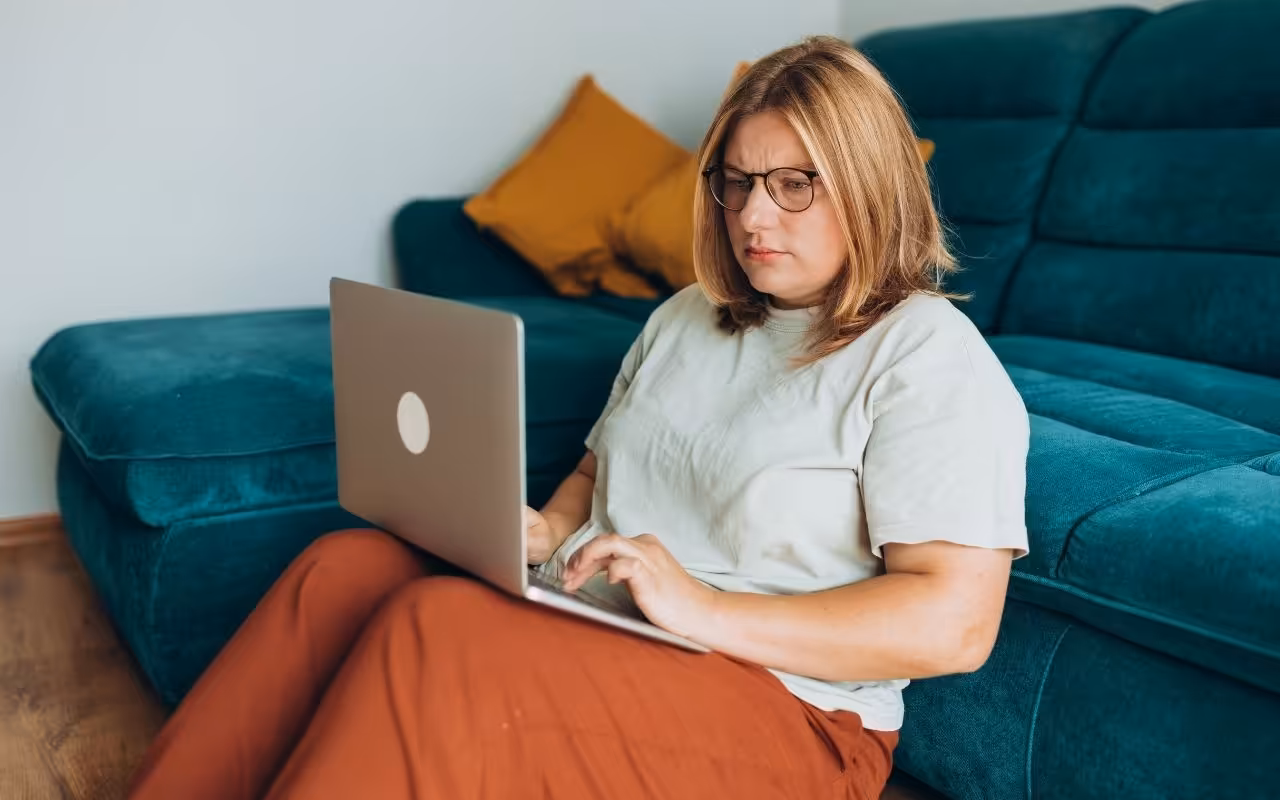 A worried woman looks closely at her laptop screen while sitting on the living room floor, wary of online job scams.